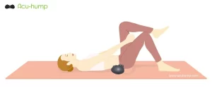 A woman is lying on a yoga mat with her legs bent, holding her left leg with her hands and pulling it towards her chest An Acu-hump is placed under the sacurm to massage and stretch the SI joint to relieve sciatica hip pain