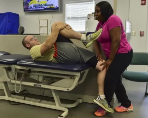 Patient with piriformis syndrome leaning on bedside stretching, holding right knee in one hand and pulling it towards chest, chiropractor helping patient to hold standing left leg